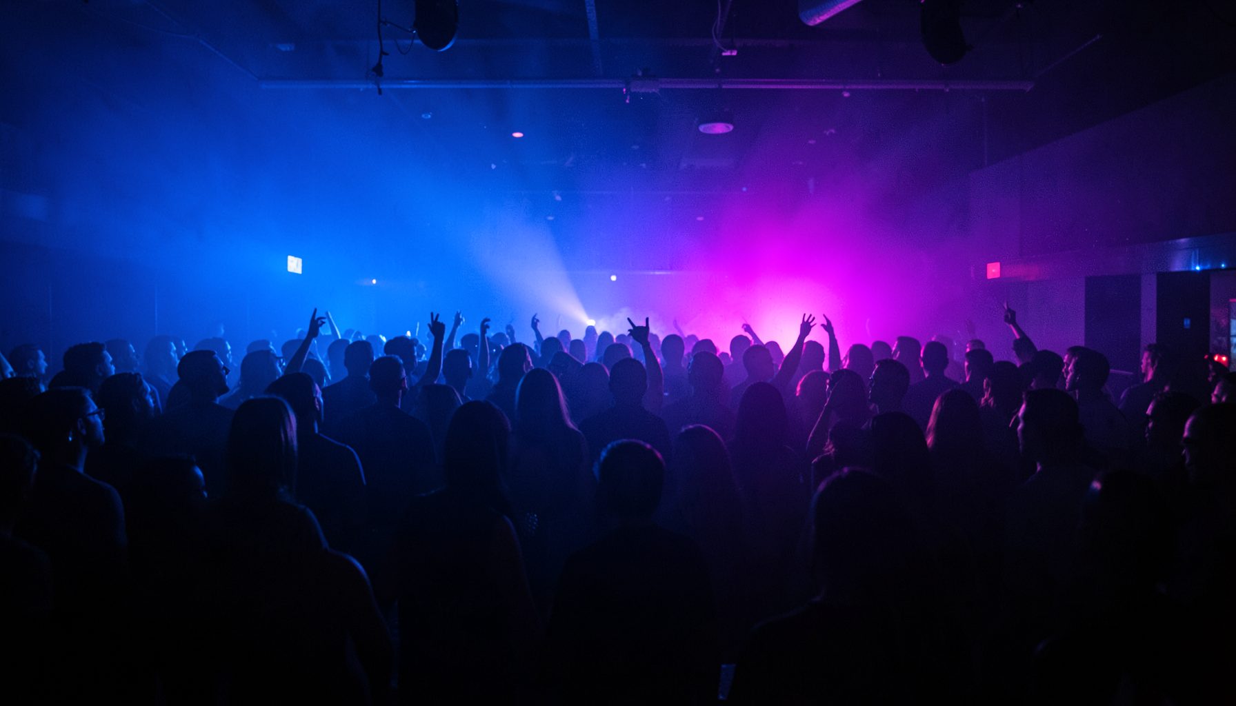 Dark nightclub crowd with hands raised, illuminated by blue and pink stage lights, energetic party atmosphere